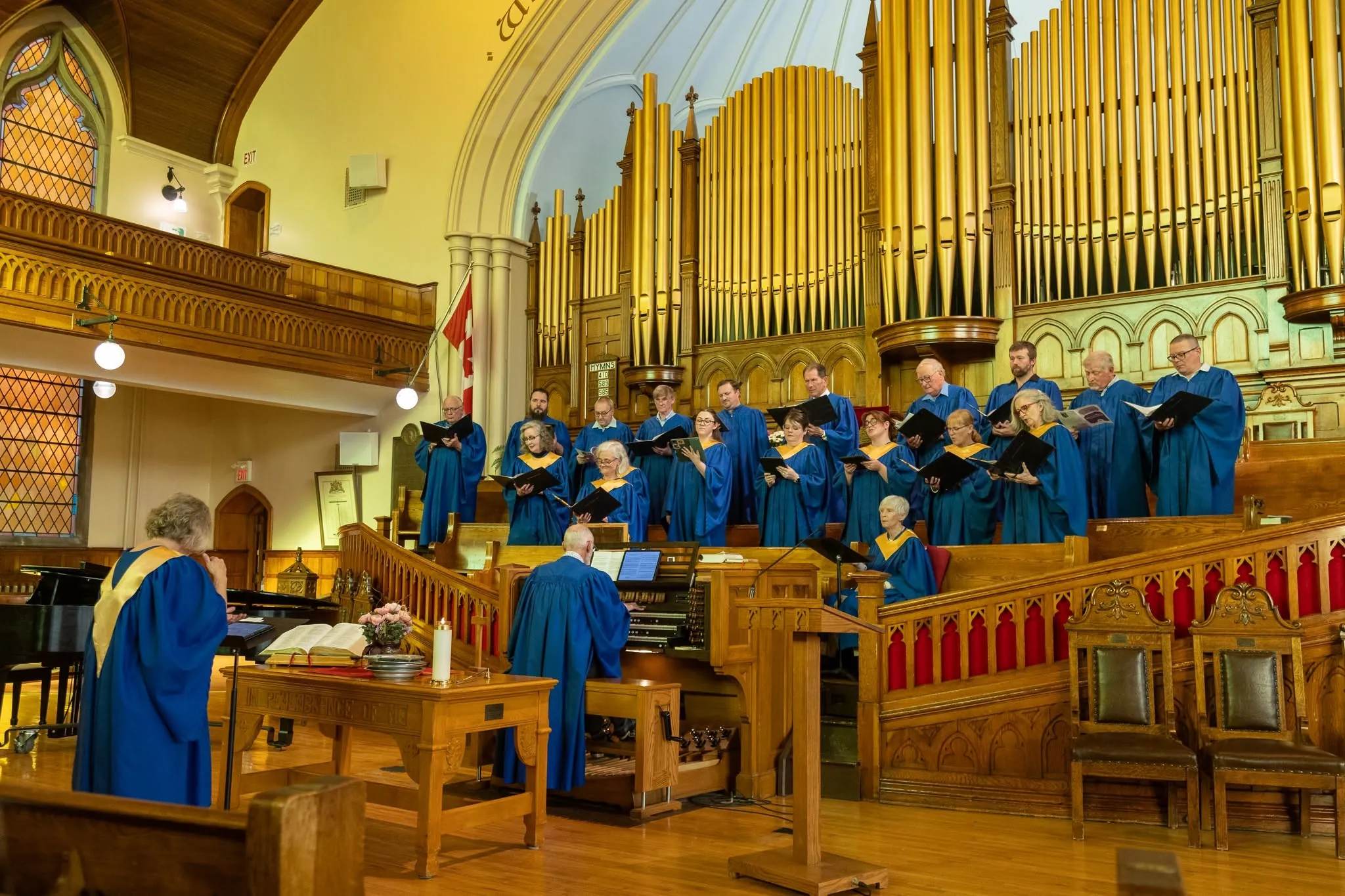 The choir in blue and gold robes singing in the chancel beneath the pipes of the 1909 Casavant organ, with the organist at the console and the director in the foreground