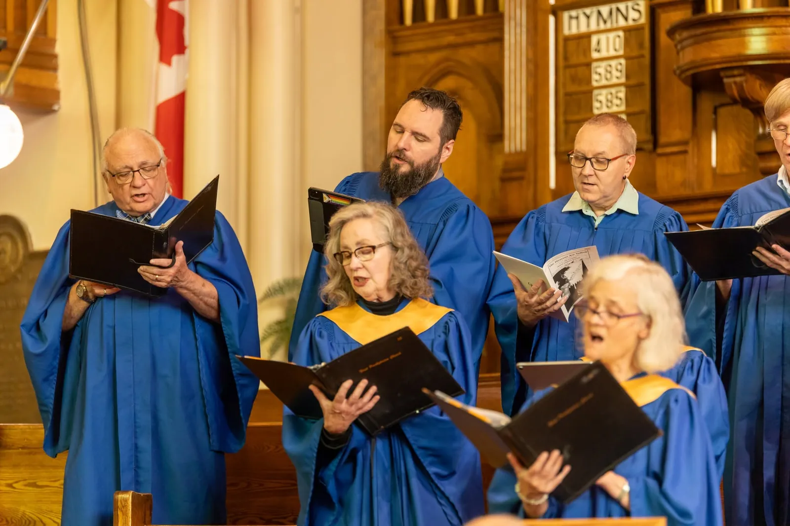 Choir members in blue and gold robes singing from their music folders, with the hymn board behind them