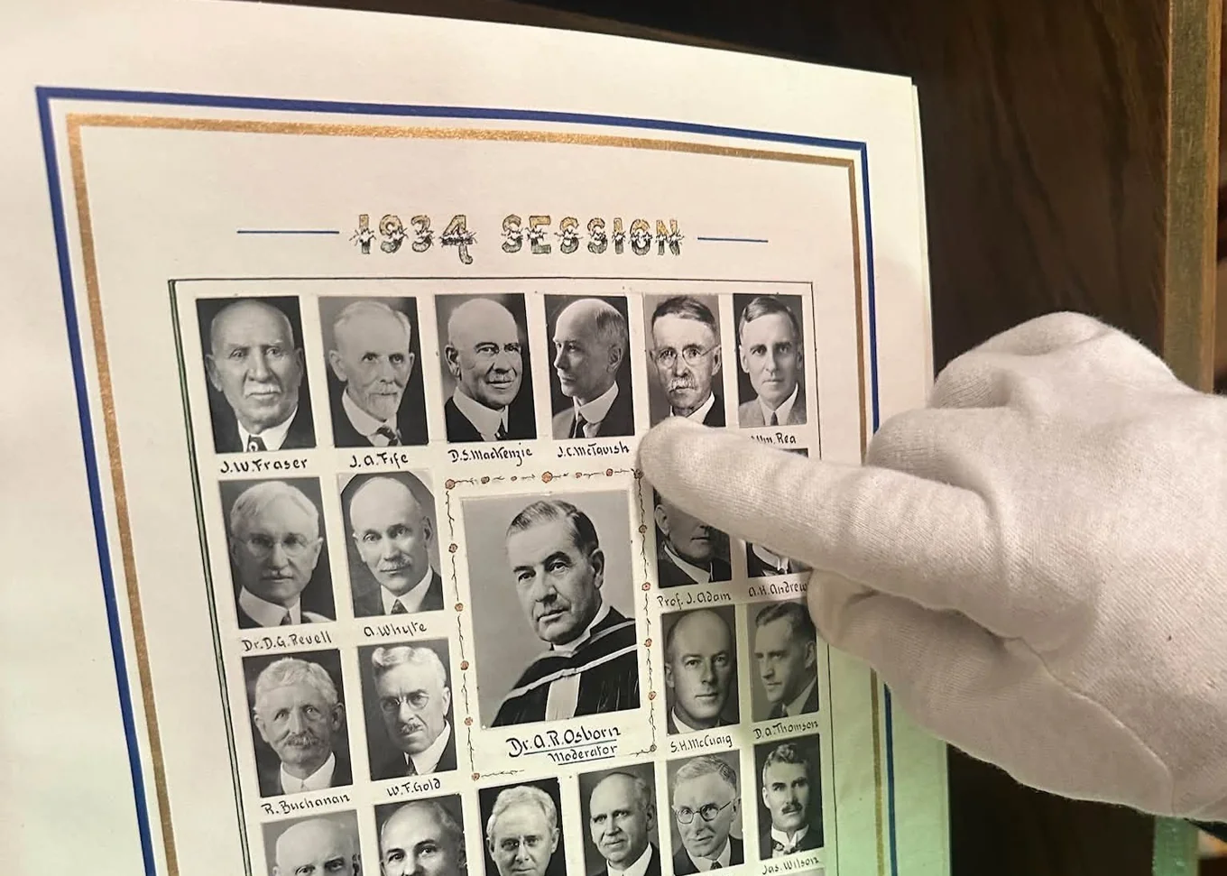 A gloved hand pointing to a portrait on a framed 1934 Session photo board